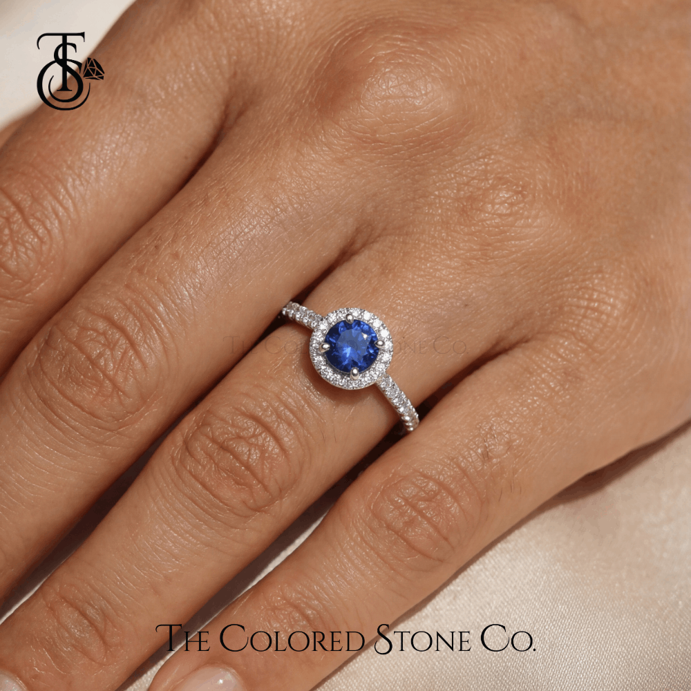 engagement Ring with blue sapphire gemstone on a hand, branded 'The Colored Stone Co'.