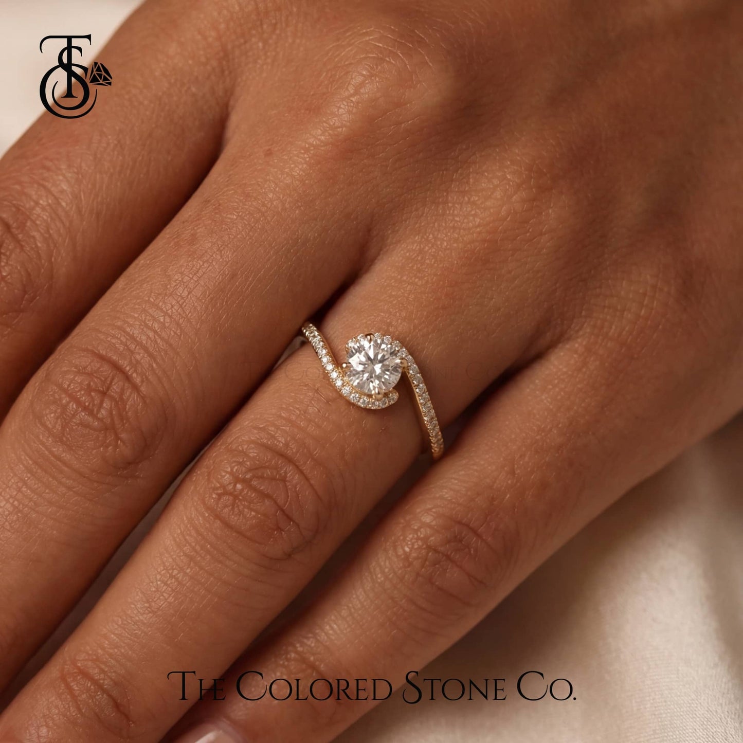 Diamond engagement ring on a finger with 'The Colored Stone Co.' branding.