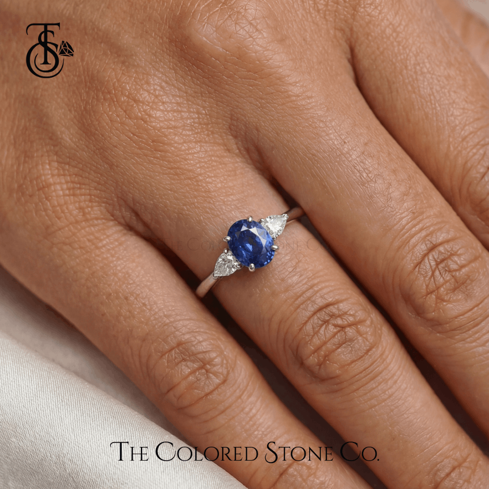 white gold engagement Ring with a blue  sapphire gemstone on a person's finger, branded 'The Colored Stone Co'.
