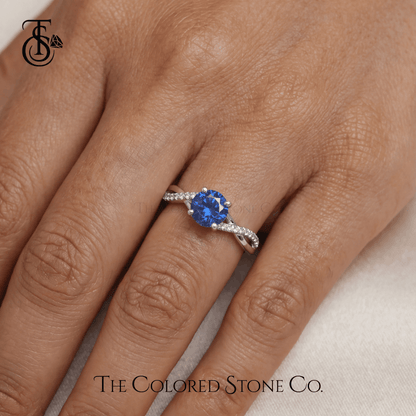 White gold engagement ring with a blue sapphire gemstone on a hand, branded 'The Colored Stone Co'.