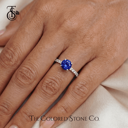 Engagement Ring with a blue sapphire gemstone on a hand, branded 'The Colored Stone Co'.