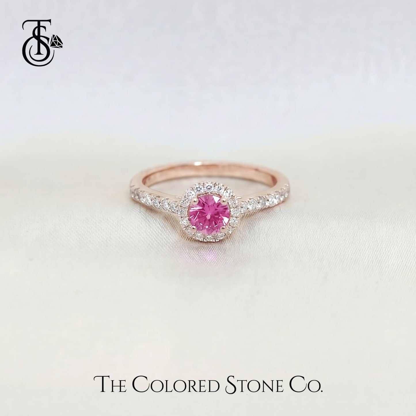 Rose gold engagement ring with a pink sapphire gemstone on a light background, branded 'The Colored Stone Co'.