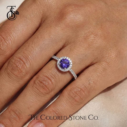 Ring with a purple gemstone on a finger, branded 'The Colored Stone Co'.