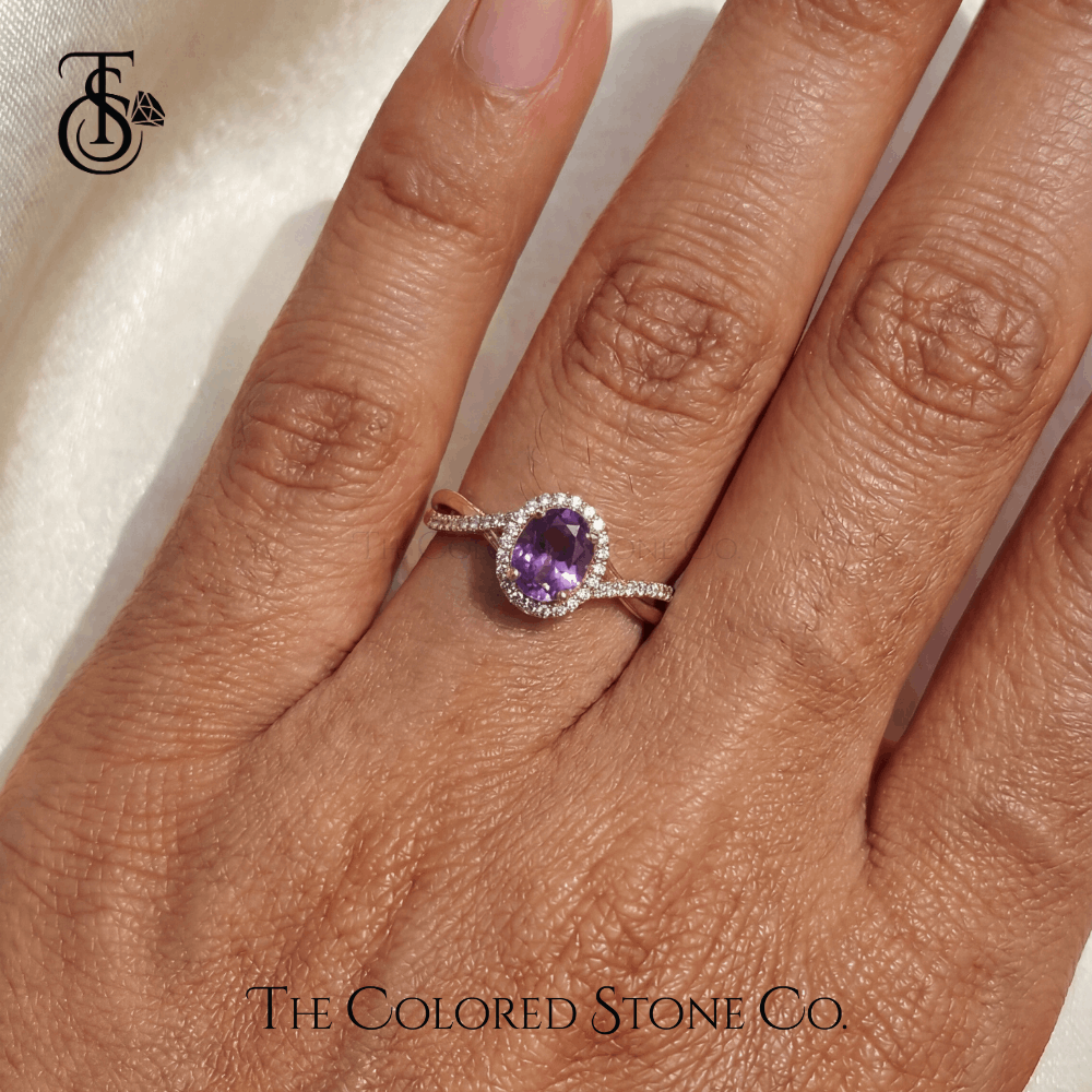 Hand wearing an engagement ring with a violet sapphire gemstone on a neutral background, featuring The Colored Stone Co. branding.