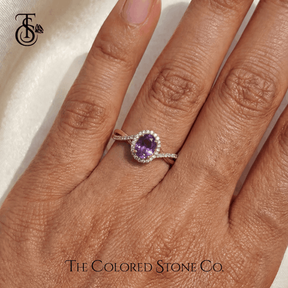 Hand wearing an engagement ring with a violet sapphire gemstone on a neutral background, featuring The Colored Stone Co. branding.