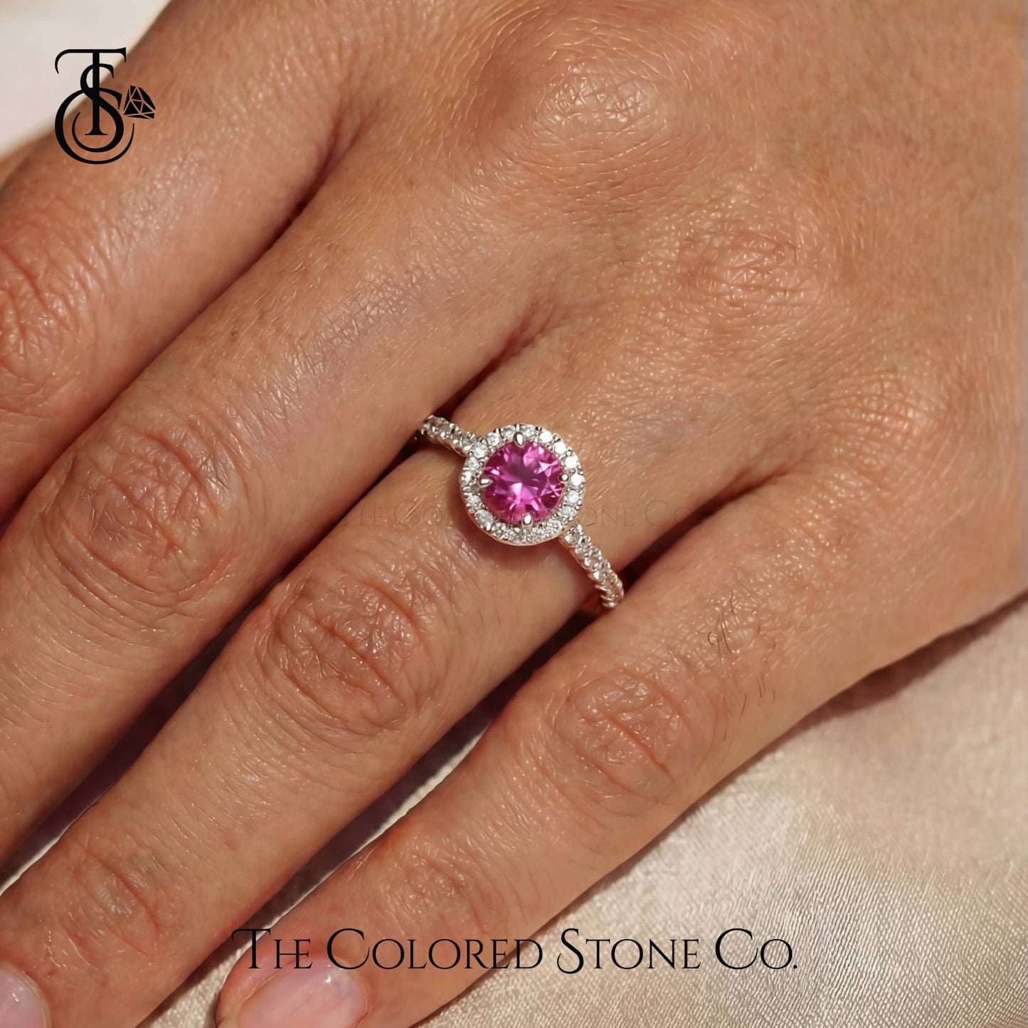Ring with a pink gemstone on a hand, branded 'The Colored Stone Co'.