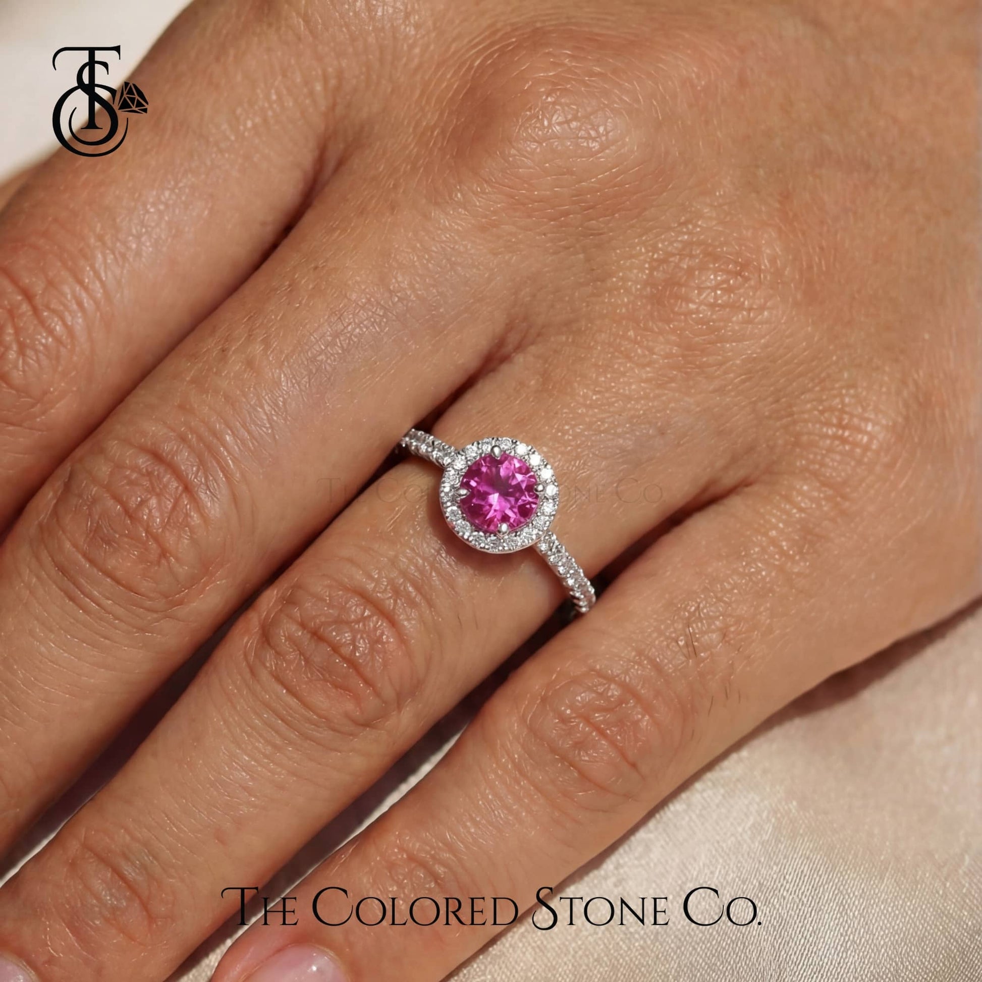 Ring with pink gemstone on a hand, branded 'The Colored Stone Co'.