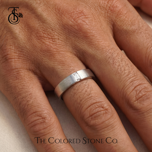 Silver men's wedding ring on a person's finger with 'The Colored Stone Co.' branding.