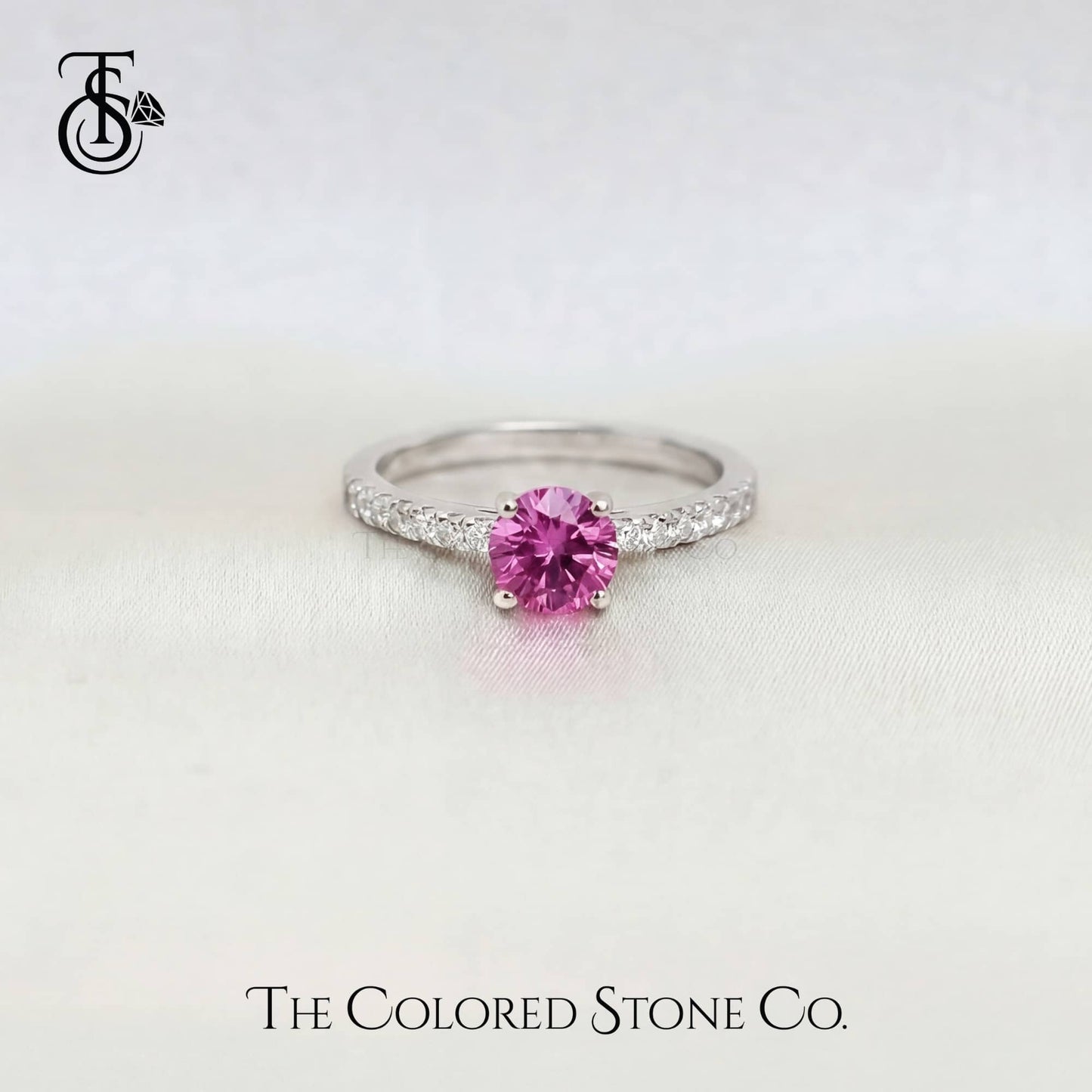 white gold engagement ring with a pink sapphire gemstone on a light gray background, featuring 'The Colored Stone Co.' branding.