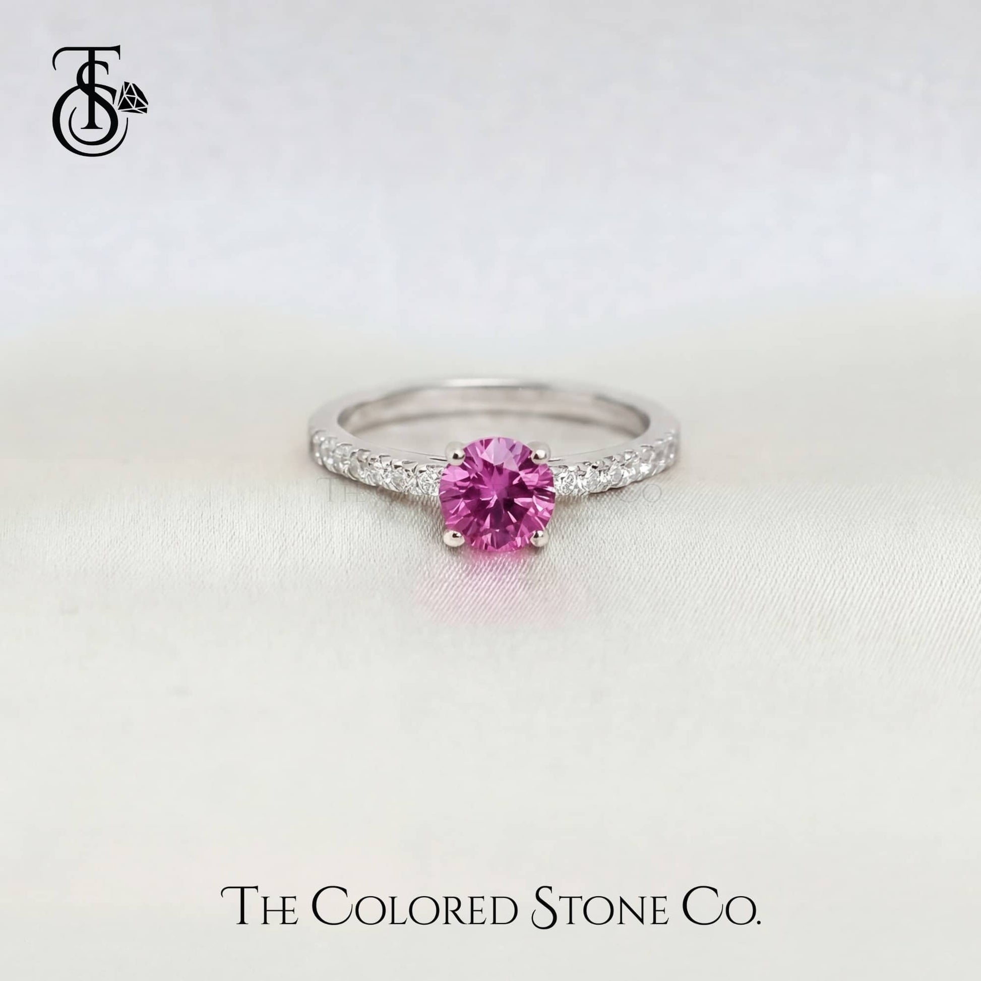 white gold engagement ring with a pink sapphire gemstone on a light gray background, featuring 'The Colored Stone Co.' branding.