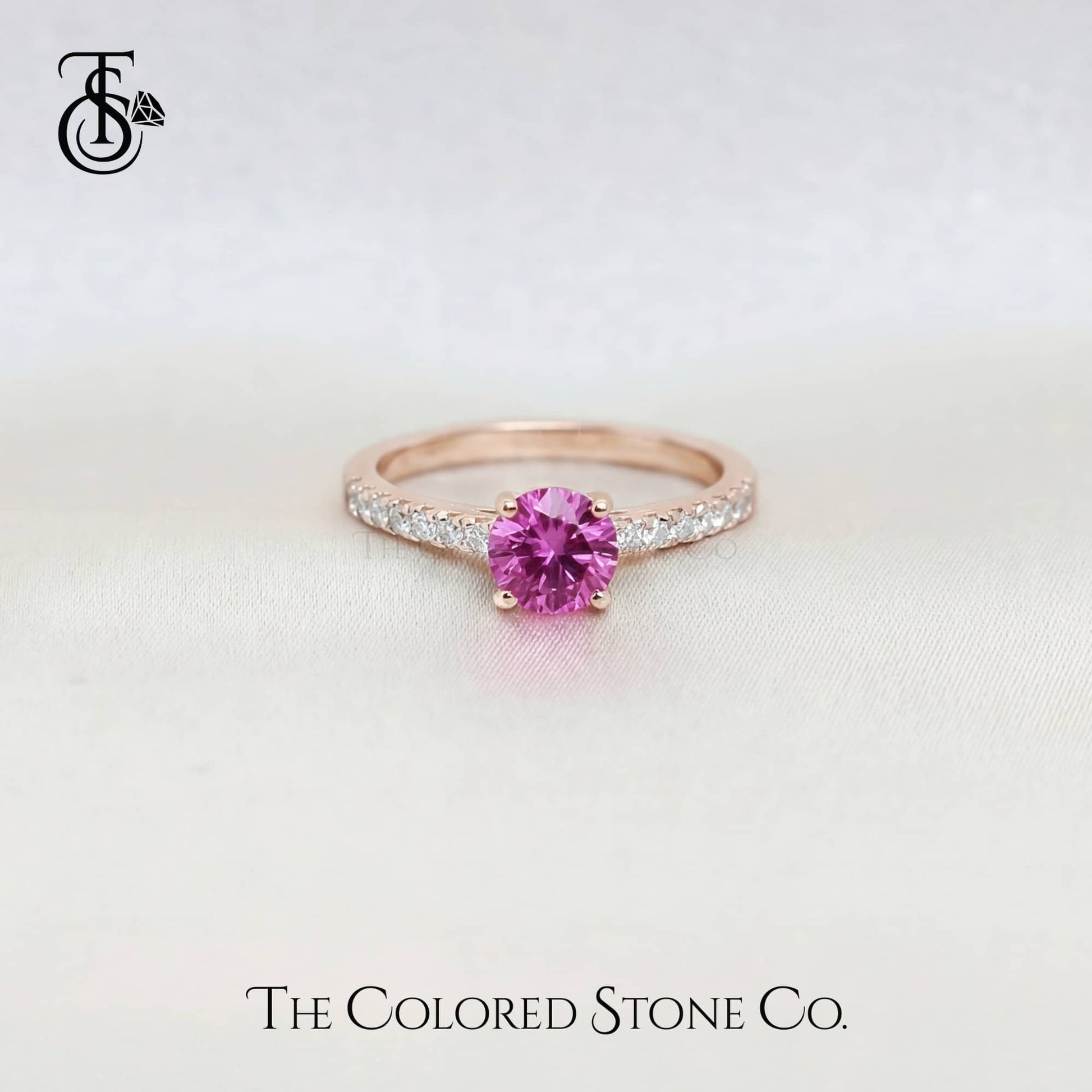 Rose gold engagement ring with a pink sapphire gemstone on a light background, branded 'The Colored Stone Co'.