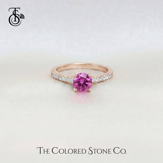 Rose gold engagement ring with a pink sapphire gemstone on a light background, branded 'The Colored Stone Co'.