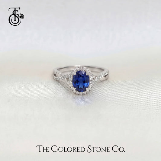 white gold engagement ring with a blue sapphire gemstone on a white background, branded 'The Colored Stone Co'.