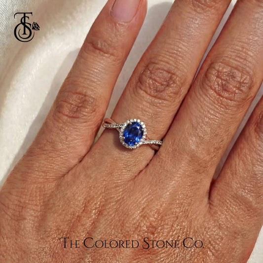 Ring with a blue gemstone on a hand, branded 'The Colored Stone Co'.
