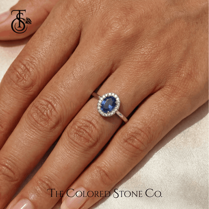 Engagement Ring with a blue sapphire gemstone on a hand, featuring The Colored Stone Co. logo.