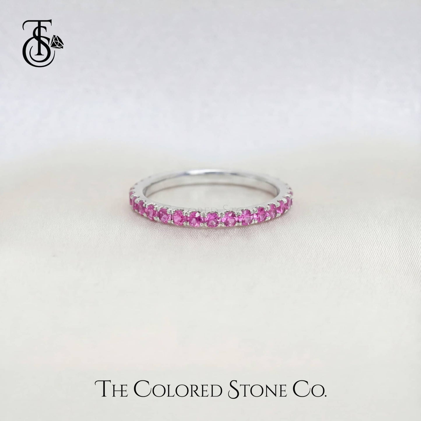 white gold wedding ring with pink stones on a white background, branded 'The Colored Stone Co'.