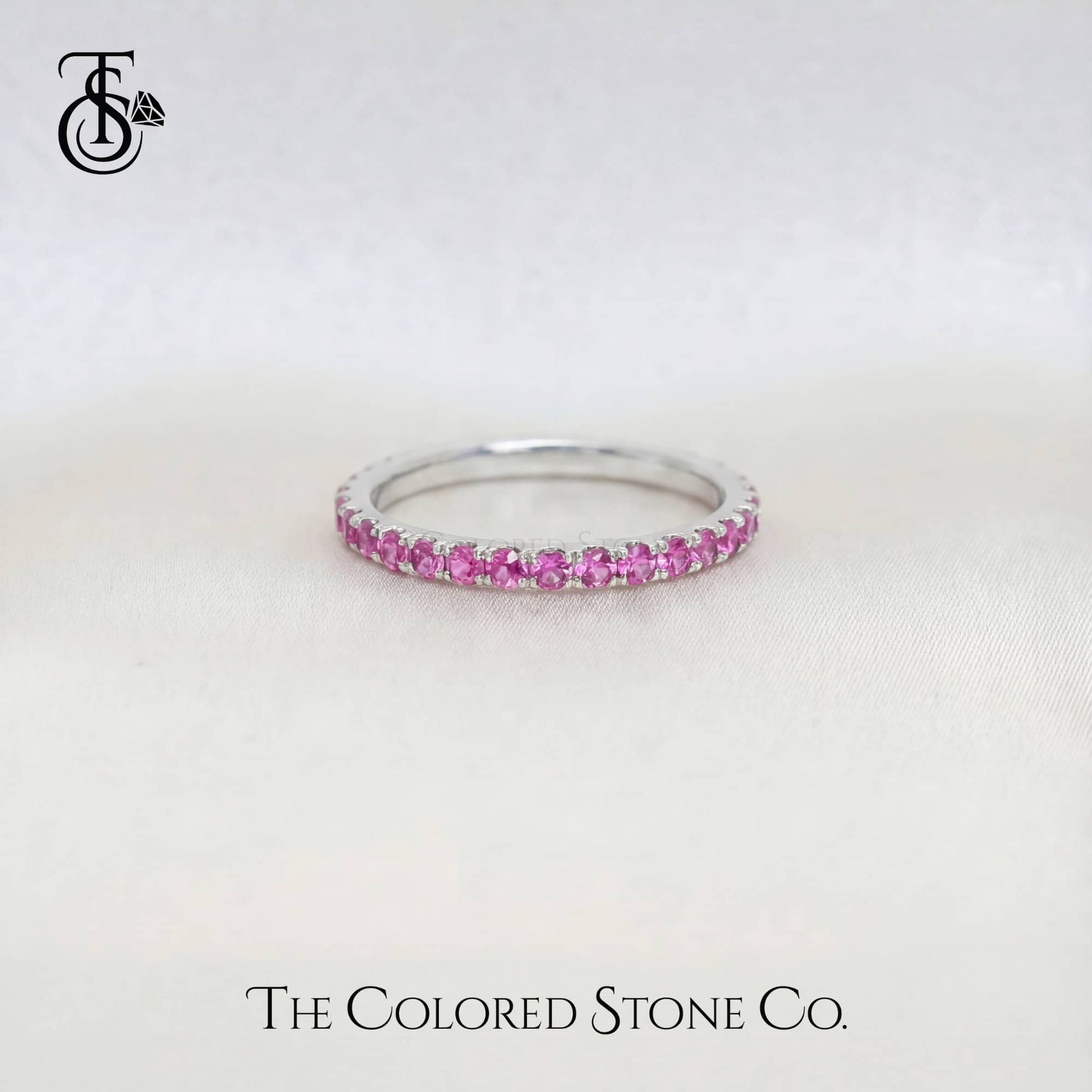 white gold wedding ring with pink stones on a white background, branded 'The Colored Stone Co'.