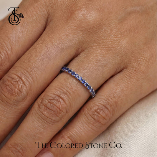 white gold wedding ring with blue stones on a finger, branded 'The Colored Stone Co'.