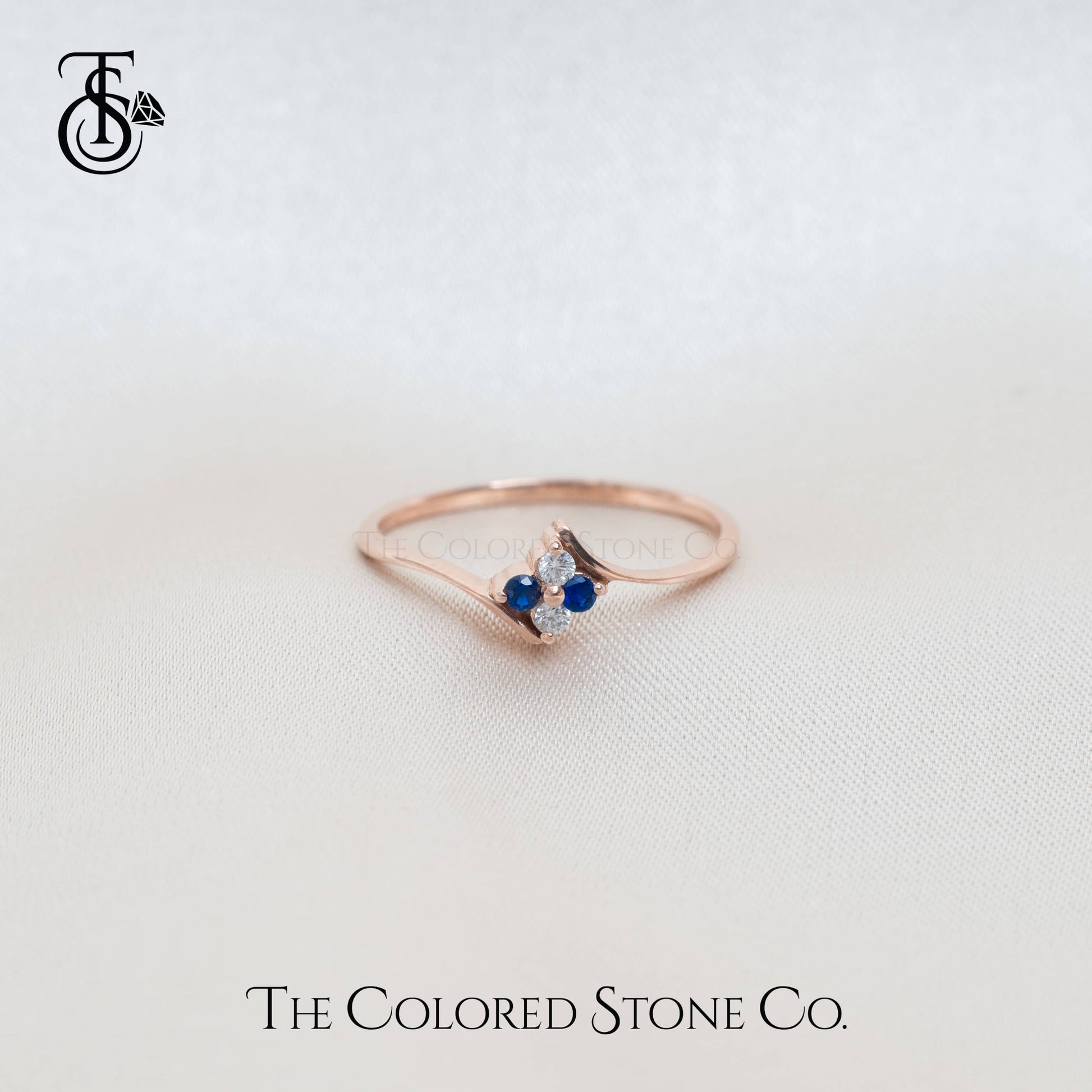 Rose gold ring with blue gemstone on a light background, featuring 'The Colored Stone Co.' branding.