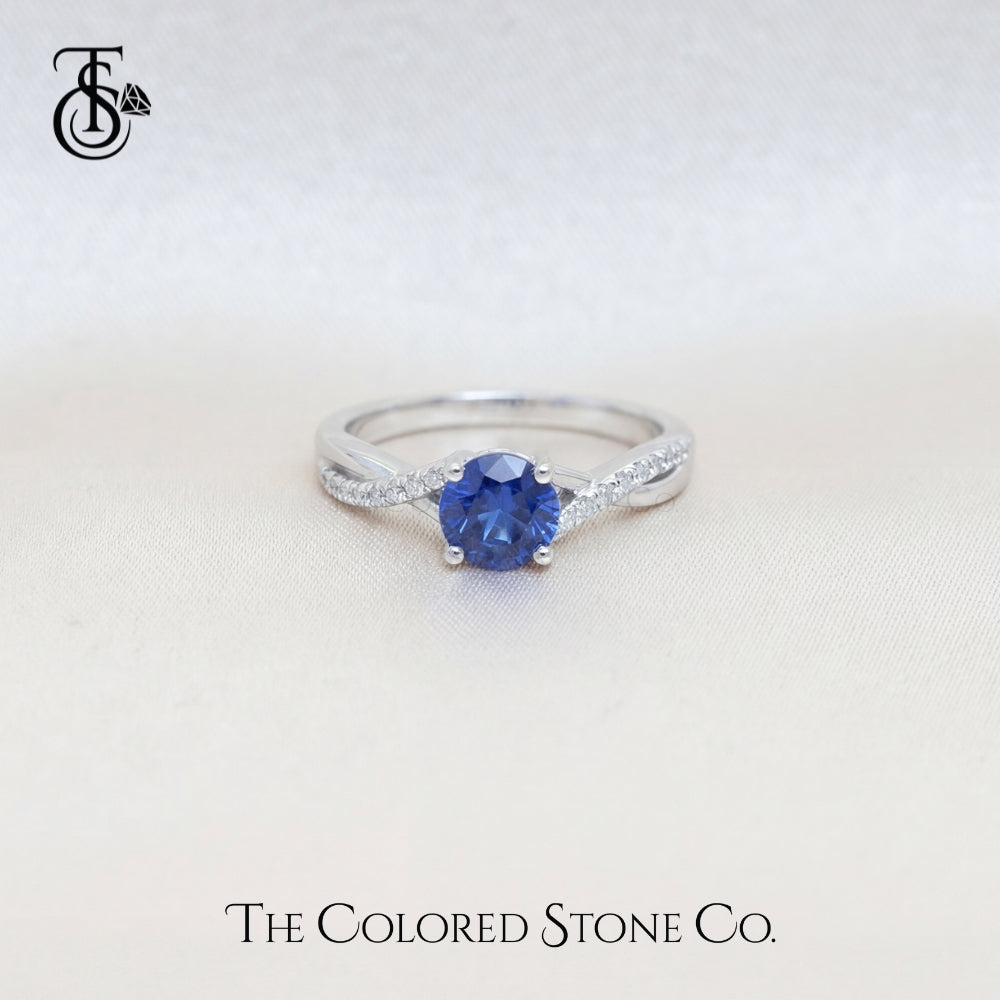 white gold engagement ring with a blue sapphire gemstone on a light background, featuring 'The Colored Stone Co.' branding.