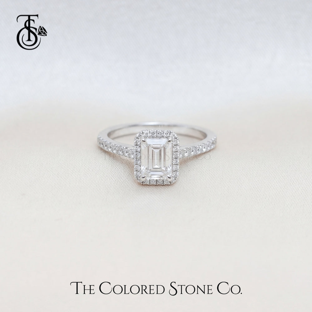 Diamond engagement ring with an emerald cut center stone on a light background, featuring The Colored Stone Co. logo.