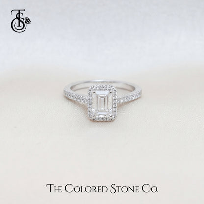 Diamond engagement ring with an emerald cut center stone on a light background, featuring The Colored Stone Co. logo.