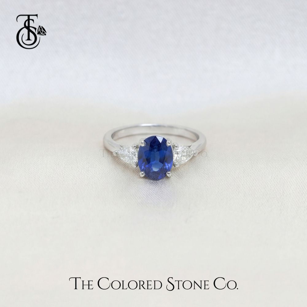 white gold engagement ring with a blue sapphire gemstone on a white background, branded 'The Colored Stone Co'.