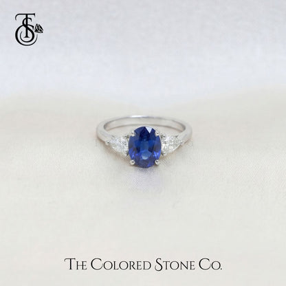 white gold engagement ring with a blue sapphire gemstone on a white background, branded 'The Colored Stone Co'.