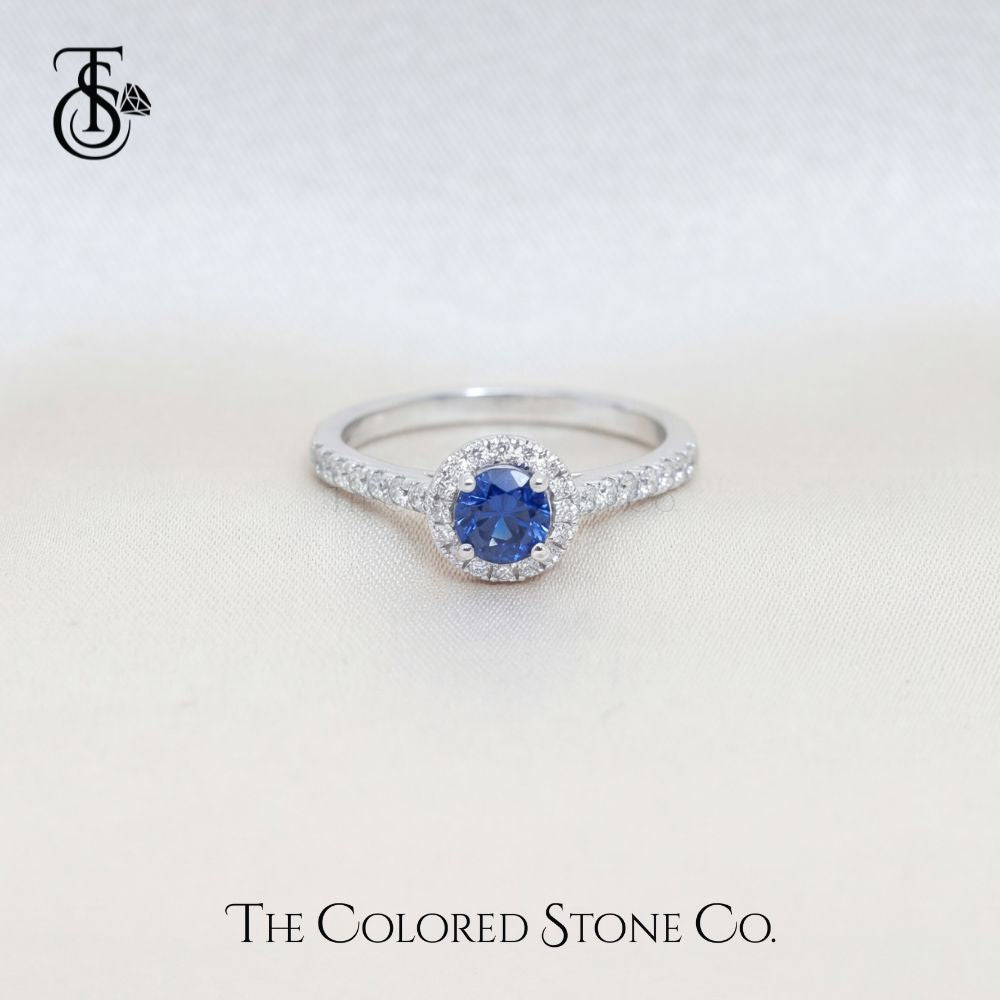 white gold engagement ring with a blue sapphire gemstone on a light gray background, featuring 'The Colored Stone Co.' branding.