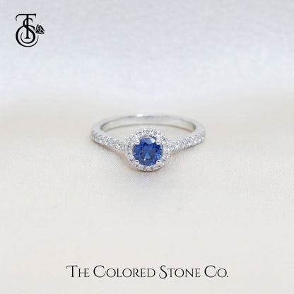 white gold engagement ring with a blue sapphire gemstone on a light gray background, featuring 'The Colored Stone Co.' branding.