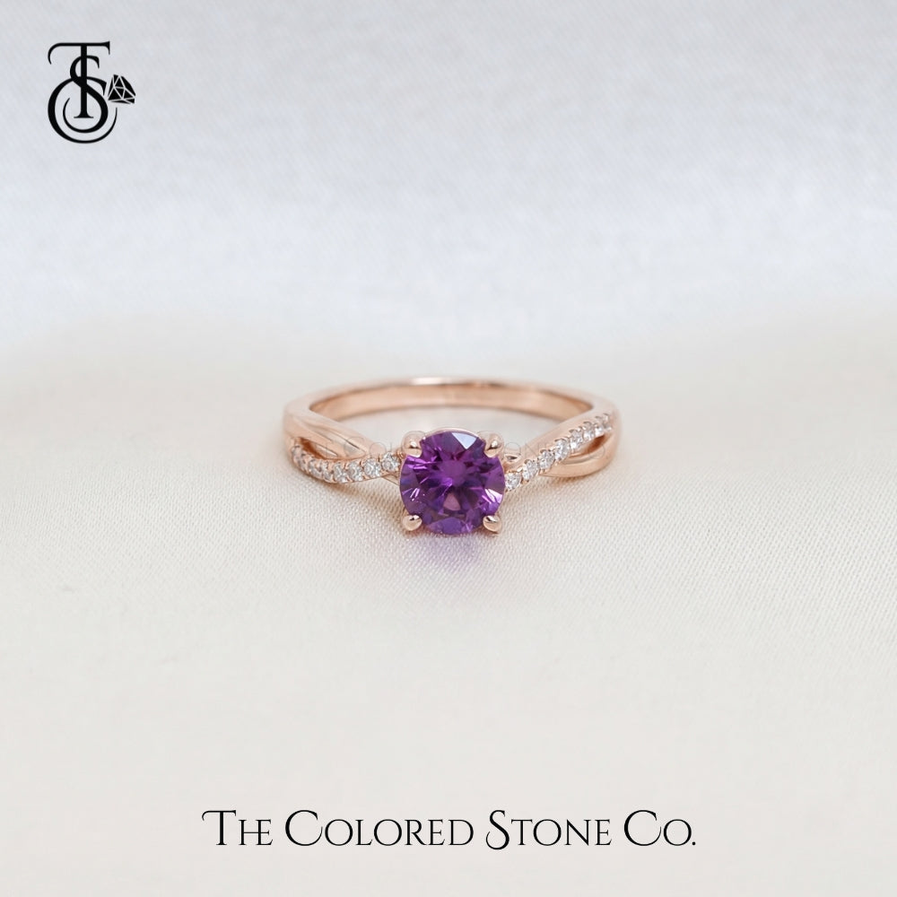 Rose gold engagement ring with a purple sapphire gemstone on a light gray background, featuring 'The Colored Stone Co.' branding.