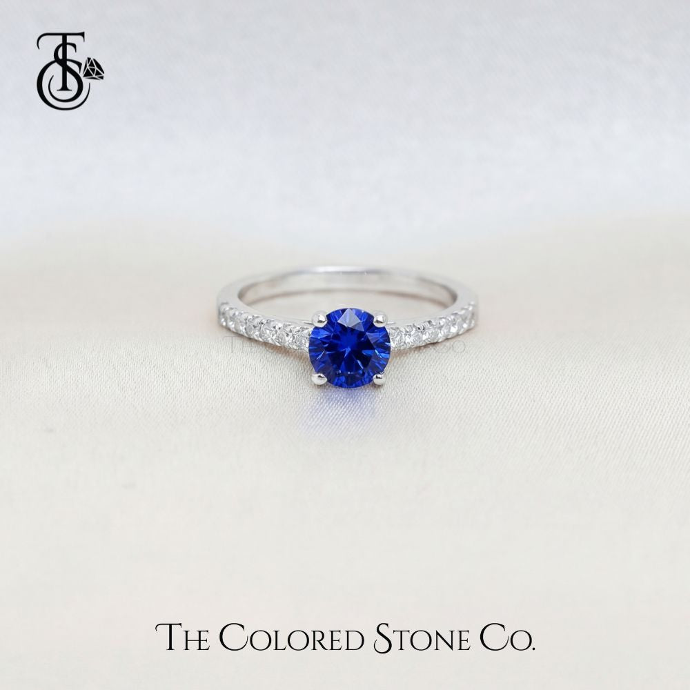 white gold engagement ring with a blue sapphire gemstone on a light gray background, featuring 'The Colored Stone Co.' branding.