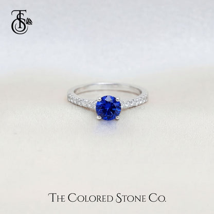 white gold engagement ring with a blue sapphire gemstone on a light gray background, featuring 'The Colored Stone Co.' branding.